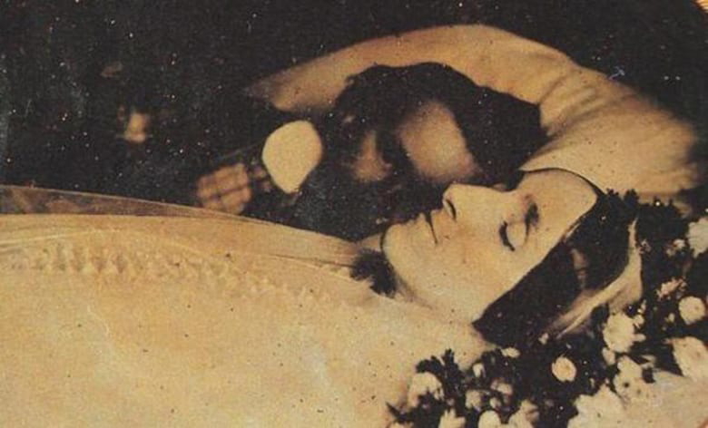 A living man mourns the death of his wife, who has been positioned surrounded by flowers and with her eyes closed. 