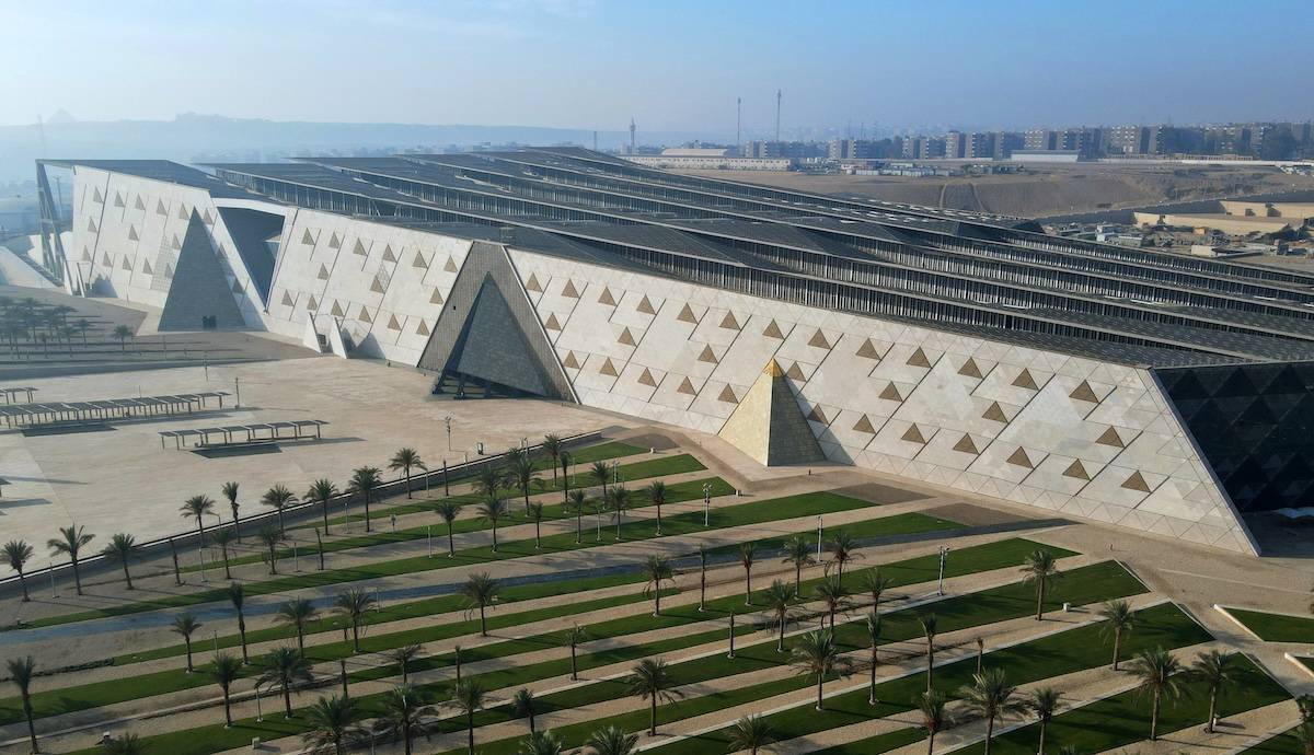 Grand Egyptian Museum exterior in Giza, showing the angular limestone façade with pyramid-shaped volumes and landscaped rows of palms; pyramids faint on the horizon.