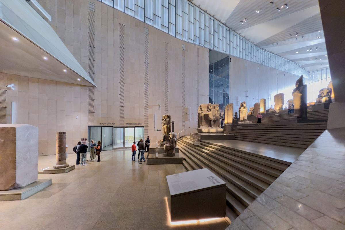 Interior of the Grand Egyptian Museum showing the Grand Staircase lined with seated pharaoh statues and stelae, visitors at the base, and daylight filtering from high windows.