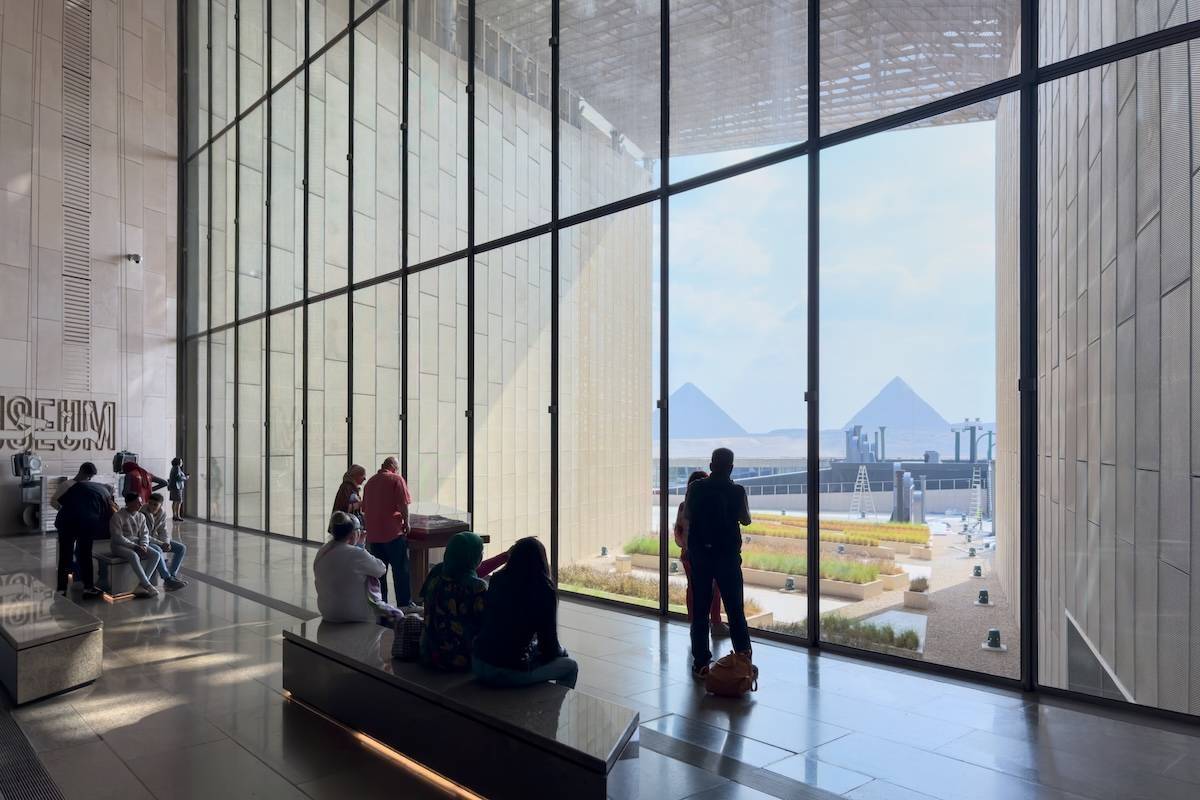Interior of the Grand Egyptian Museum with a tall glass wall framing two Giza pyramids; visitors sit and stand in the atrium overlooking the landscaped courtyard.